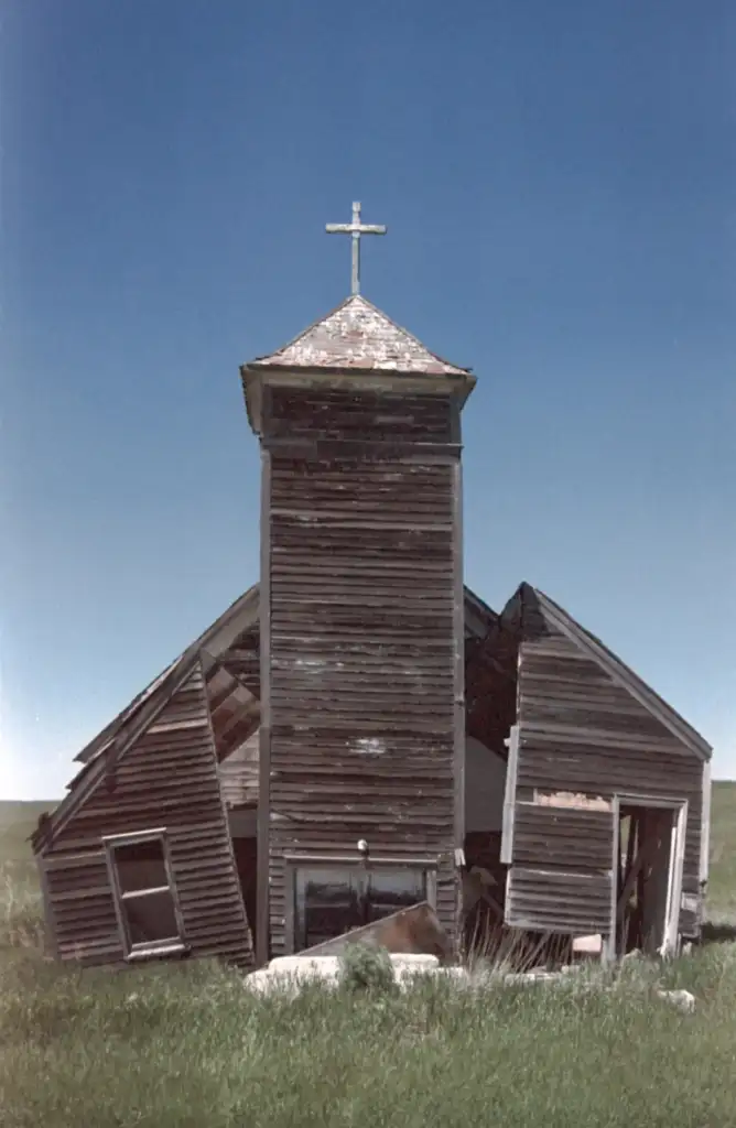 A dilapidated wooden building with a cross on top, resembling a church, sits in a grassy field. The structure leans to one side, with visible broken windows and a partially collapsed roof against a clear blue sky.