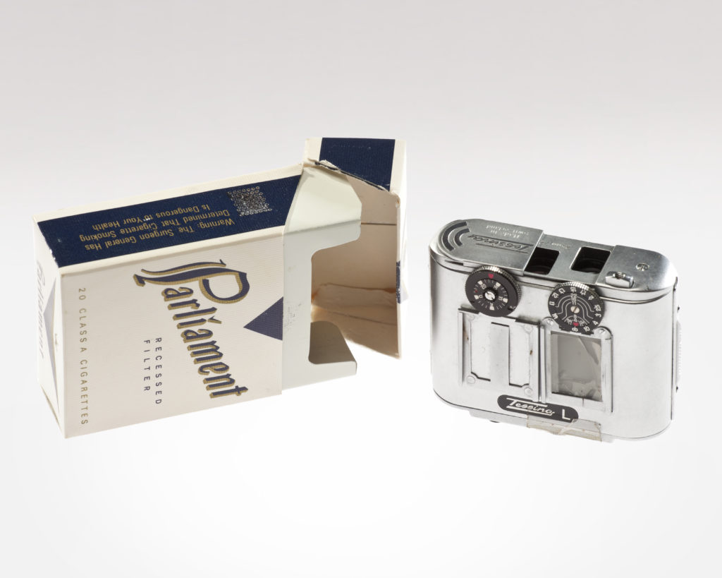 A vintage Zeiss Ikon camera next to an open Parliament cigarette pack. The cigarette pack is partially torn and lying on its side, showcasing its retro design. The camera features two dials and a shiny metallic body.