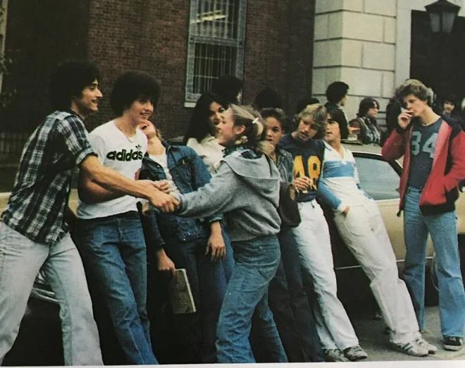 A group of teenagers stands casually in front of a building and a parked car. They are engaged in conversation and appear relaxed. One girl is playfully holding two boys by their jackets, as others look on, smiling or talking.