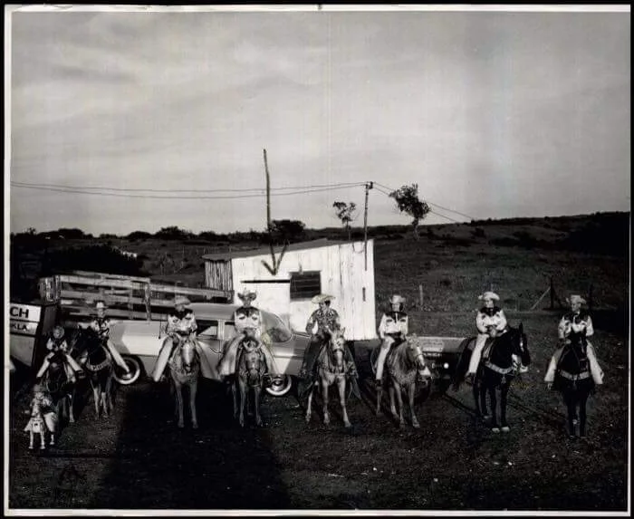 A group of people wearing western attire sit on horseback, with children and dogs alongside them. They are arranged in front of a small rustic building with a pickup truck parked nearby in an open field. The sky is overcast.