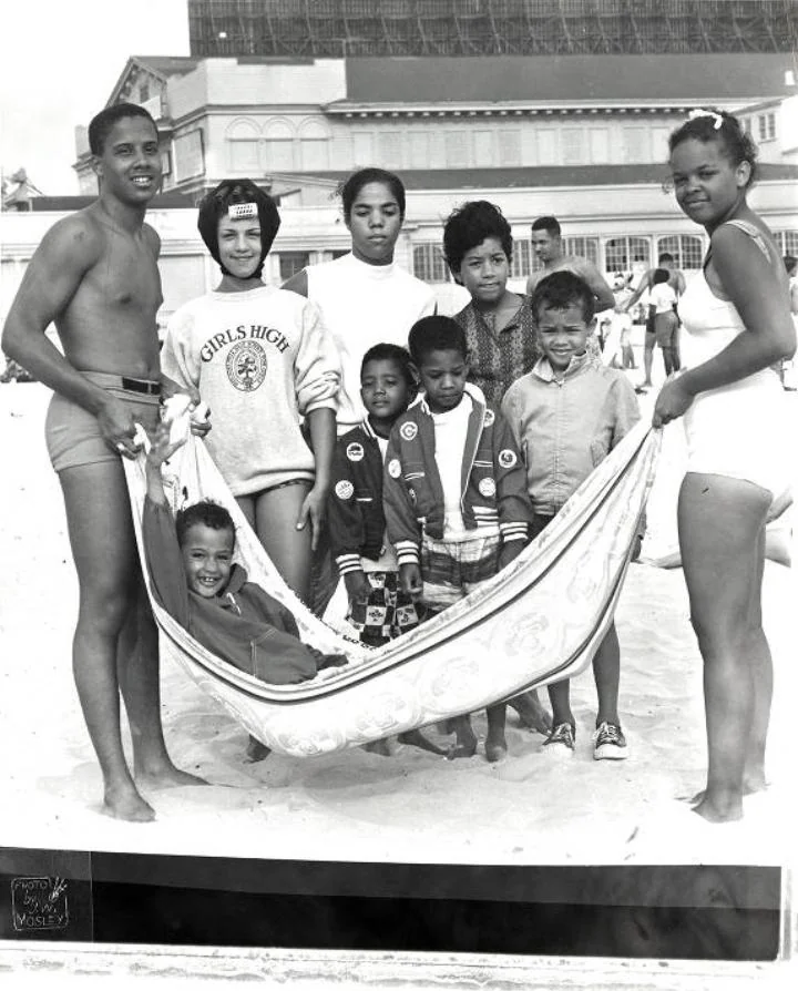 A group of people on a beach, with two adults holding a cloth hammock carrying a smiling child. Behind them, five children stand closely together. The background features a large building and other beachgoers. The scene is in black and white.