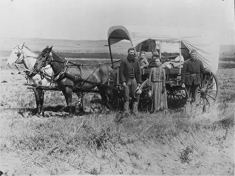 A black and white photo showing four people posing beside a covered wagon pulled by two horses. The scene is set in an open grassland. The individuals are dressed in 19th-century attire, and various supplies are visible in the wagon.