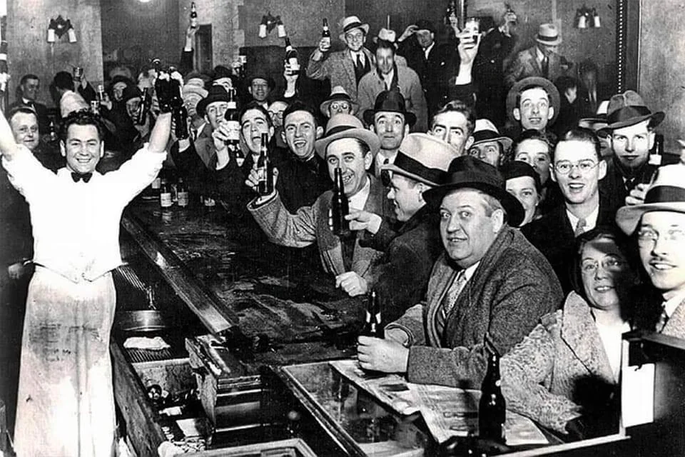 A crowded bar scene in a vintage black-and-white photo featuring a bartender with raised arms and a large, cheerful crowd of men and women holding up bottles and glasses, seemingly celebrating or toasting. They are dressed in 1920s-1930s style clothing.