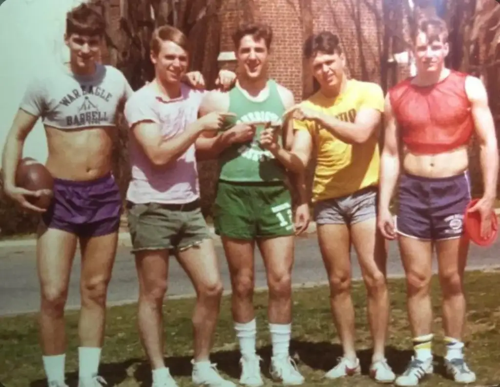 Five young men in casual, sporty outfits stand outdoors on grass. Two point towards one in a green tank top. Others wear shorts and t-shirts, one holding a football. A brick building and road are in the background.