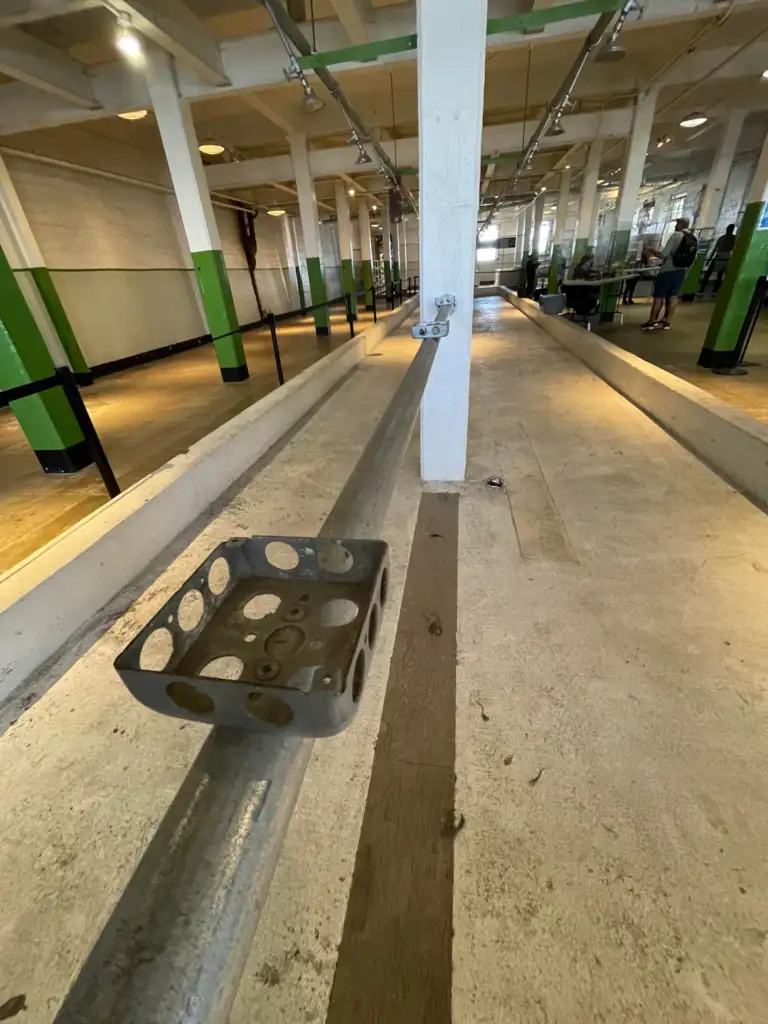 A long indoor bowling alley with a metal bowling pin setter on a track in the foreground. The floor is wooden, and green and white columns line the sides. The space is spacious, with muted lighting.