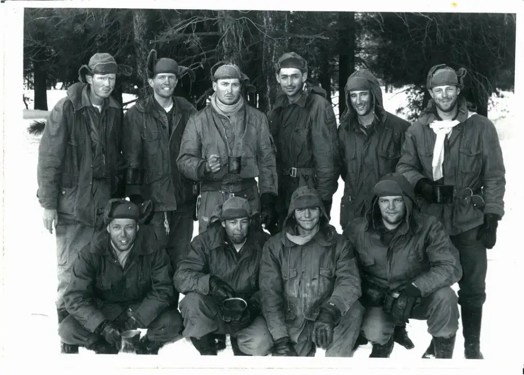 A group of ten men dressed in cold weather gear and hats pose for a black and white photo. Some hold mugs, and they stand and kneel on snow-covered ground, with trees in the background.