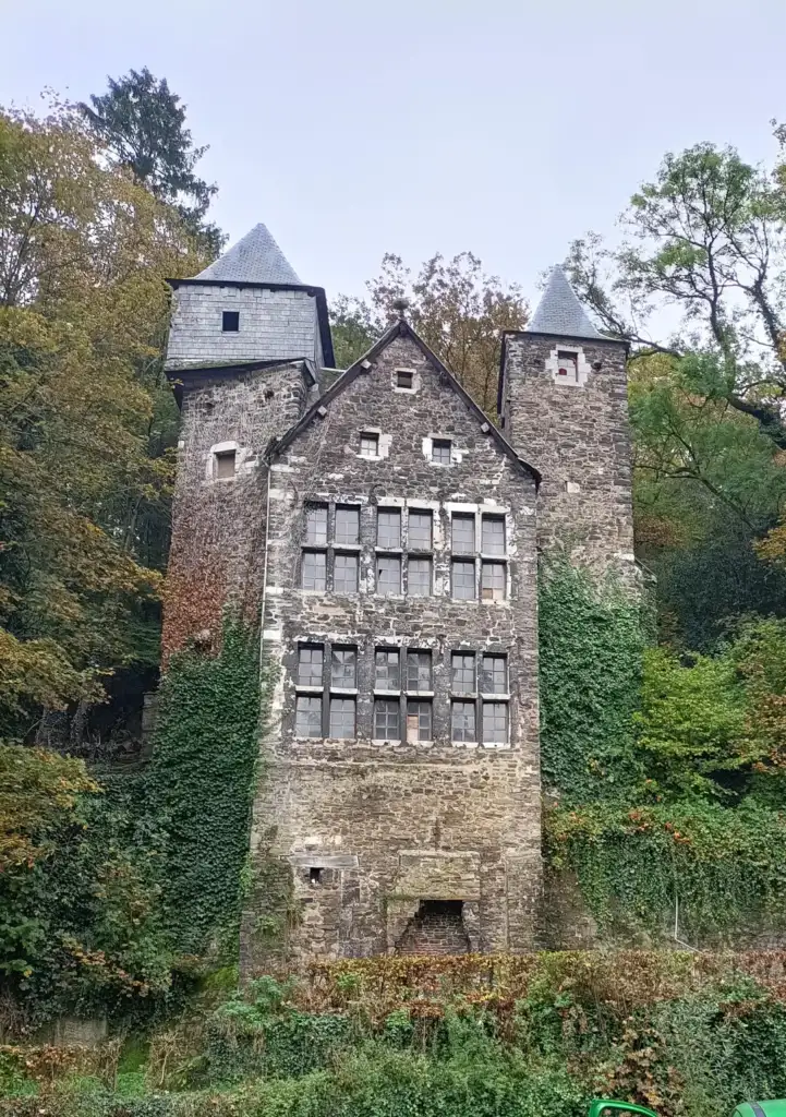 A rustic stone building with steep gabled roofs, surrounded by dense greenery and trees. The facade features multiple windows, and ivy climbs the walls, giving it an old, abandoned appearance. The structure is set against a backdrop of lush foliage.