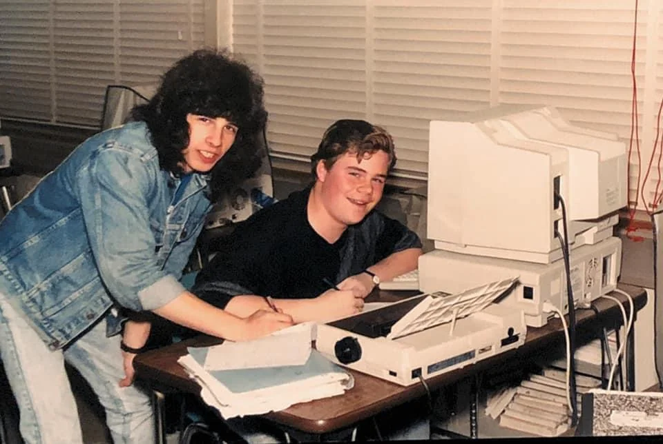 Two young people are sitting at a desk with vintage computer equipment. One person with long hair wears a denim jacket and leans over to write on a paper. The other sits smiling, looking at the camera. The room is filled with blinds and fluorescent lighting.