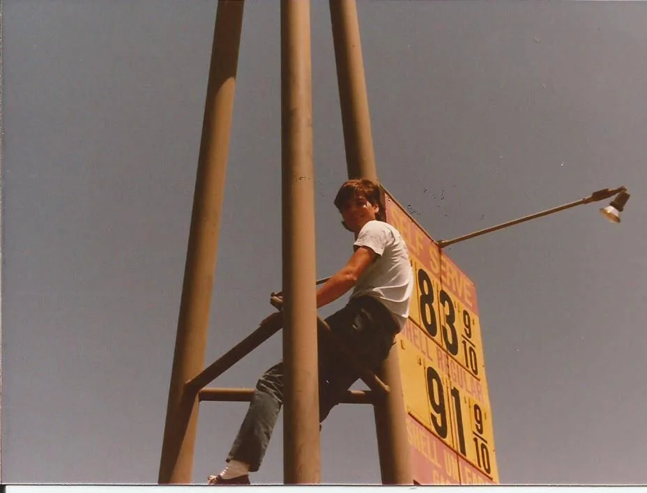 A person climbs a tall metal structure to change prices on a large sign displaying gas prices: 83 and 91 cents per gallon. The person is wearing a white shirt and jeans, and the sky is clear.