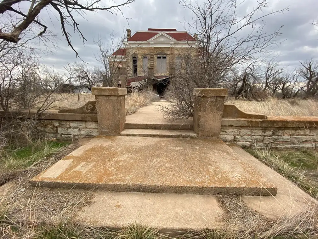 Deserted building with a grand, weathered facade, surrounded by leafless trees. A stone path leads to the entrance, flanked by two stone pillars. Overcast sky enhances the scene's eerie atmosphere. Sparse grass covers the foreground.