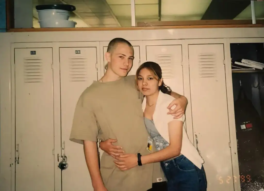 Two teenagers pose in front of lockers. The boy, with a shaved head and wearing a beige T-shirt, embraces the girl with brown hair, wearing a white shirt and jeans. The setting appears to be a school hallway.