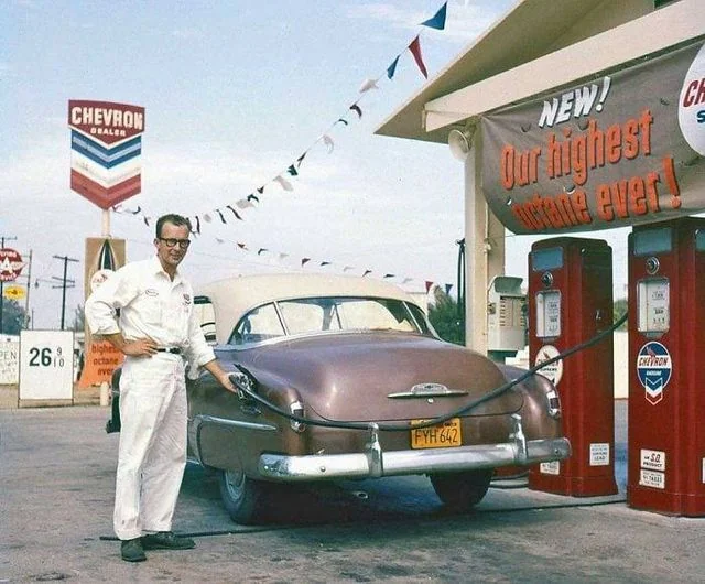 A vintage scene at a Chevron gas station features a man in a white jumpsuit refueling a classic brown car. A banner reads, "NEW! Our highest octane ever!" The gas price is shown as 26.9 cents. Colorful pennants hang above.