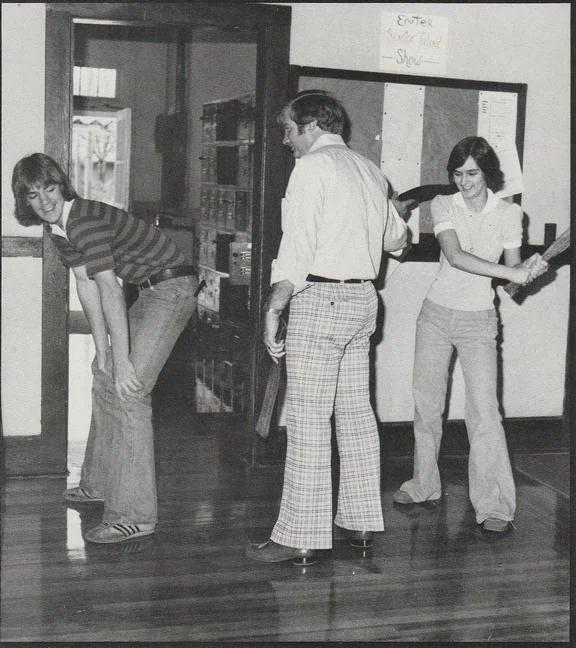 A black and white photo of three people indoors. One person bends over playfully while a man in plaid pants stands between two others. A person on the right pretends to paddle the bending person. They all appear to be having fun.