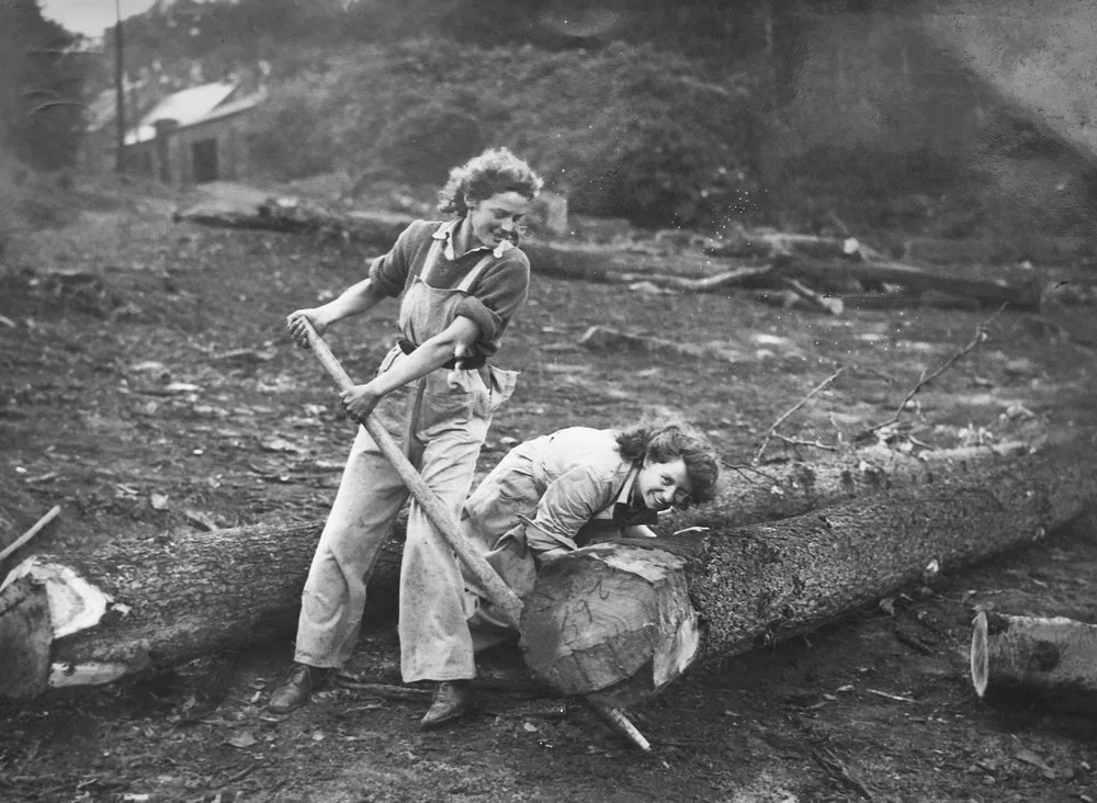 Two women in work overalls use a two-person saw to cut through a large log outdoors. One stands pulling the saw, while the other leans over the log, both appearing focused and determined. The background features trees and a small building.