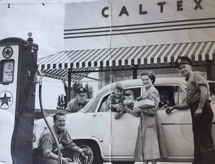 A black-and-white vintage photo showing a family at a Caltex gas station. Two attendants are servicing the car: one fueling up and another checking a tire. A woman stands beside the car holding papers, and two children lean out the window smiling.