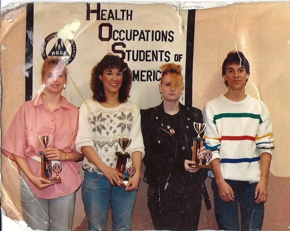 Four young adults stand in front of a "Health Occupations Students of America" banner, each holding a trophy. The group includes three women and one man, dressed casually. One woman has a punk style with a unique hairstyle.