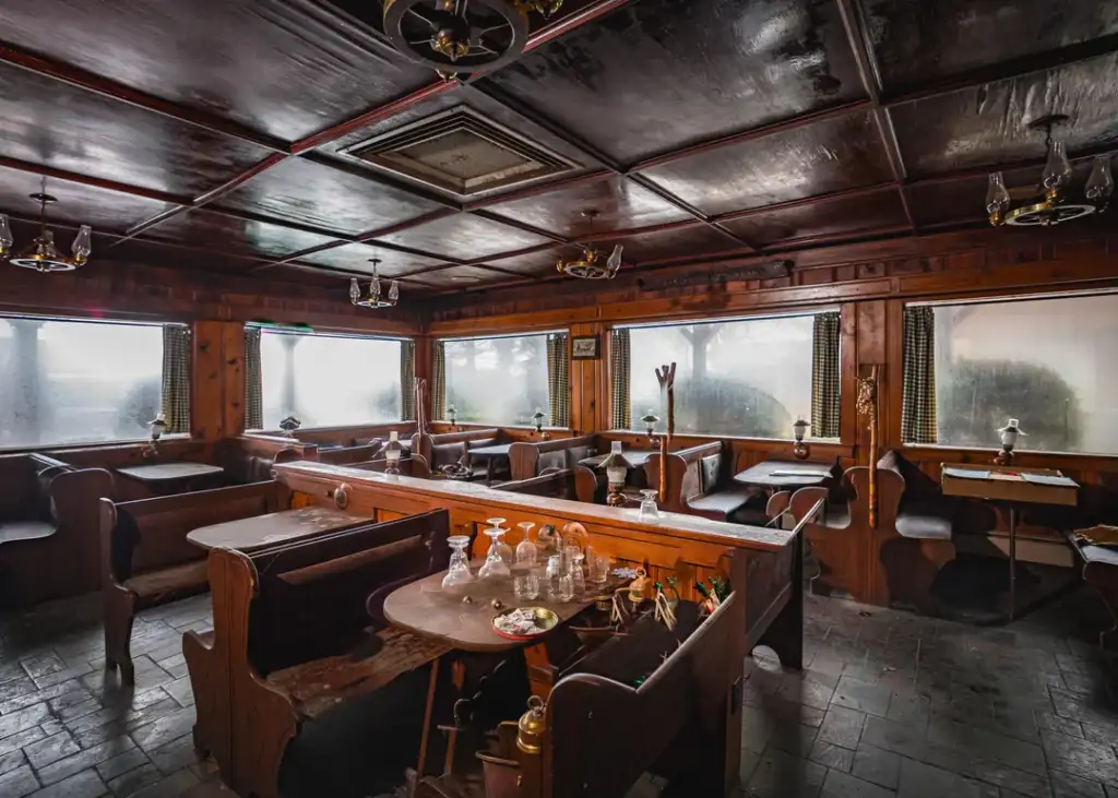 A dimly lit, rustic wooden restaurant interior with empty tables and booth seating. Several glasses and candlesticks are on the tables. Large windows with frosted glass line the walls, creating a cozy atmosphere.