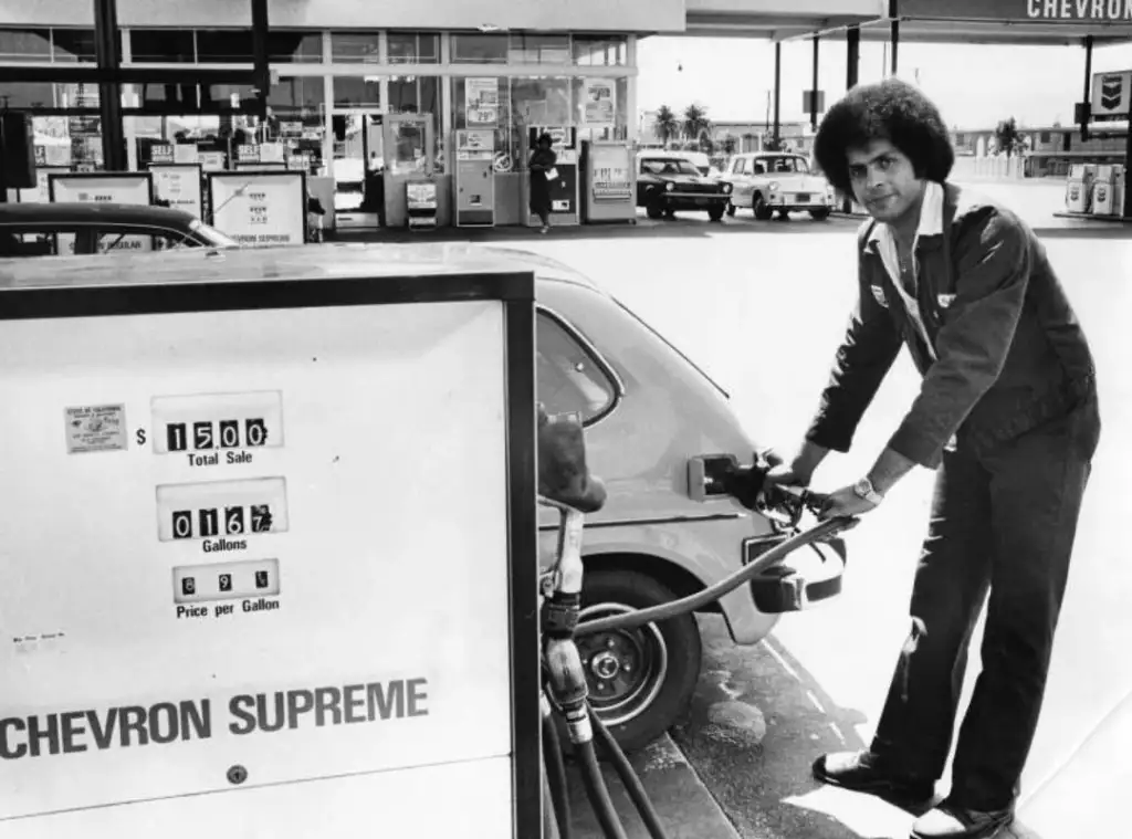 A person with an Afro hairstyle is seen pumping gas into a small car at a Chevron station. The gas pump shows $15.00 total sale, 0.08 gallons, and $2.64 price per gallon. The background features more pumps and a Chevron sign. The image is in black and white.