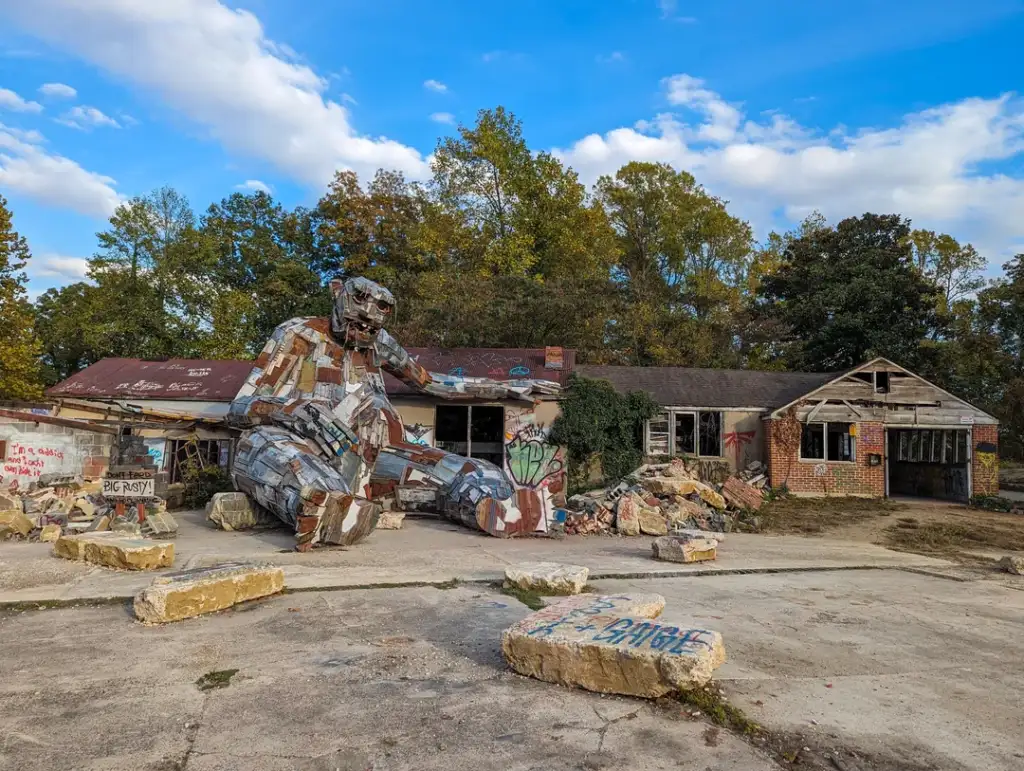 A giant sculpture made from metal and recycled materials sits in front of an abandoned, graffiti-covered building. The structure resembles a reclining figure. Surrounding are trees with autumn foliage under a blue sky with scattered clouds.