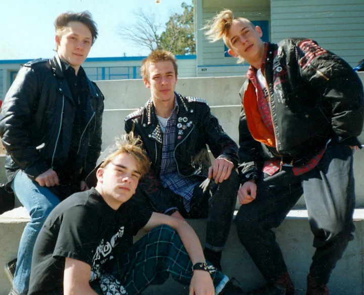 Four young men pose on outdoor steps, wearing punk attire. They sport leather jackets, plaid pants, and spiked or styled hair. The background features a light-colored building and a blue sky.