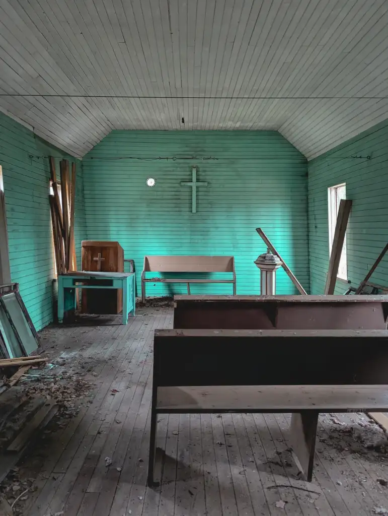 An abandoned church with teal-painted wooden interior. A cross hangs on the back wall. Pews, a pulpit, and debris are scattered on the dusty wooden floor. Dim natural light enters through windows on the sides.