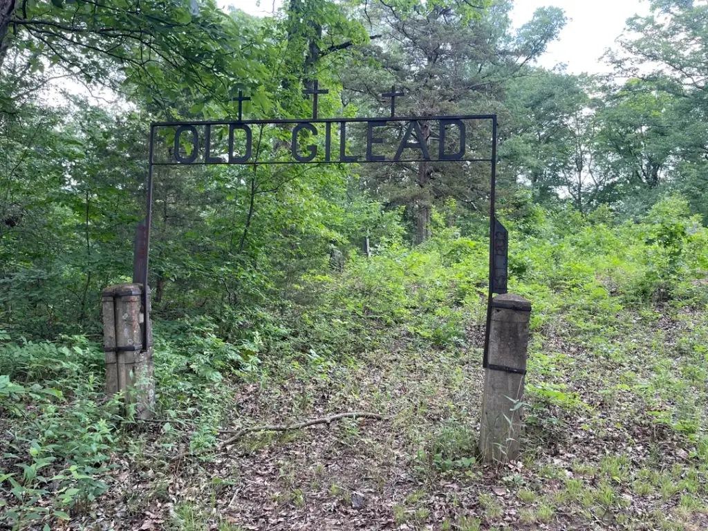 A weathered metal archway with the text "OLD GILEAD" stands between two stone pillars in a lush, green forest. Dense trees and undergrowth surround the area, creating a secluded and serene atmosphere.