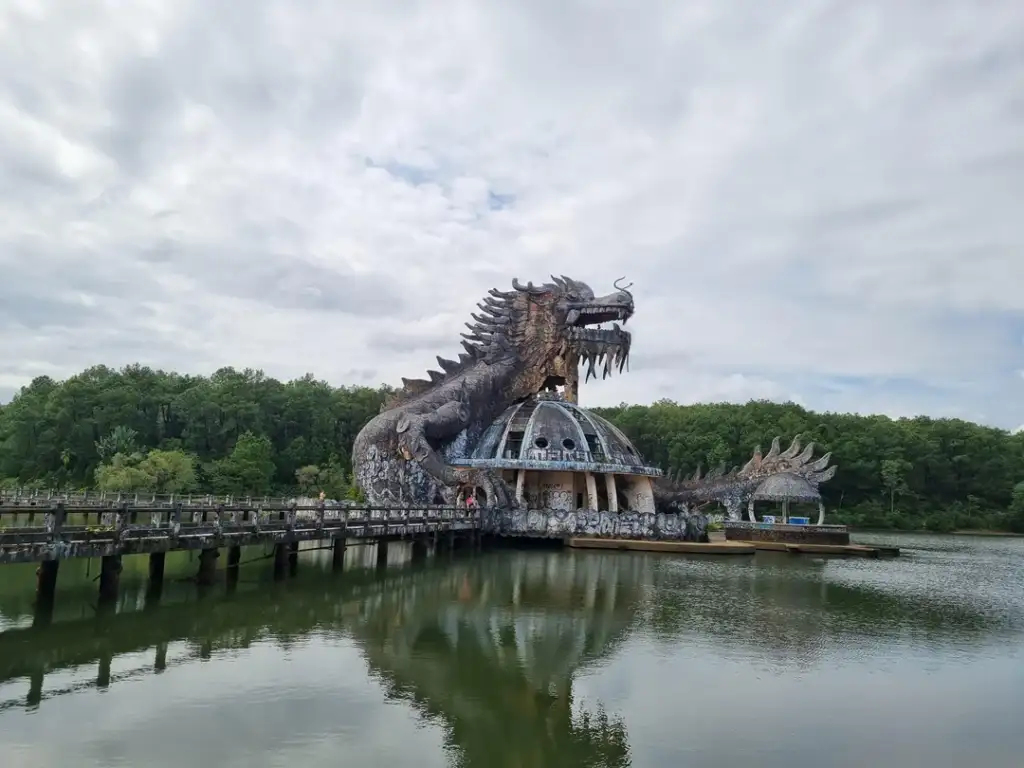 A large dragon sculpture looms over a circular building on a lake, surrounded by trees. A long walkway extends over the water to the structure. The sky is cloudy, adding a dramatic effect to the scene.