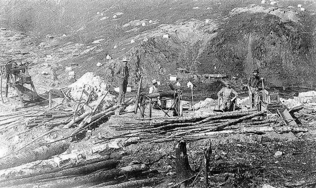 Black and white photo of four miners working on a hillside during the 19th-century gold rush. They are using partially constructed wooden structures to aid in their work. Hills and scattered rocks surround them.