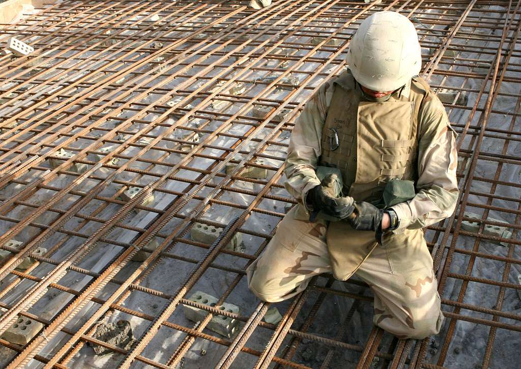 A person in military gear is kneeling and working with tied steel reinforcing bars on a construction site. The individual is wearing a helmet, a vest, and gloves, focusing on securing the rebar grid.