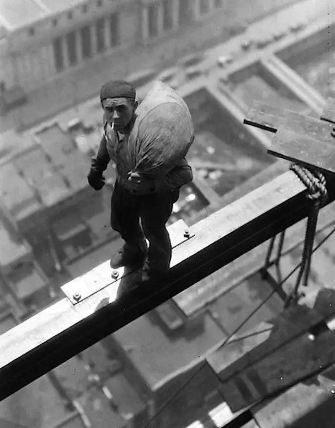 Historic black-and-white photo of a construction worker balancing on a narrow steel beam high above a city. He's carrying a large sack on his back and wears a cap and gloves. Buildings and streets are visible far below.