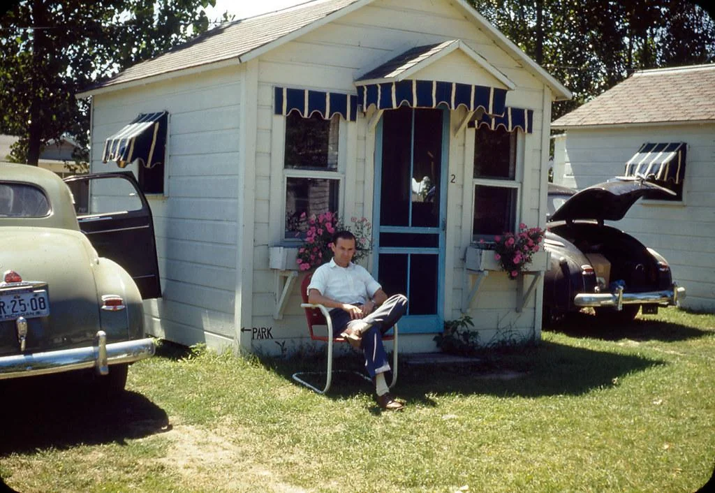 A man sits in a chair outside a small white cabin with blue trim, surrounded by two vintage cars with open doors. Pink flowers are in window boxes, and the sun casts shadows on the grassy area.