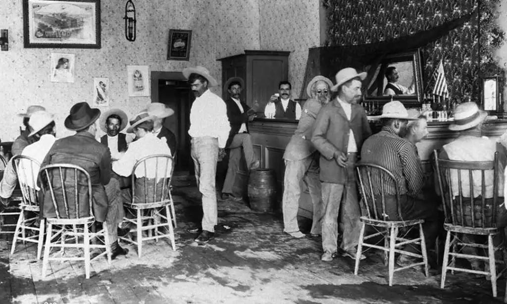 A black-and-white photo of a historical saloon with several men wearing hats sitting and standing around tables and at the bar. The room has patterned wallpaper, a mirror, and a U.S. flag in the corner. Some men are engaging in conversation.