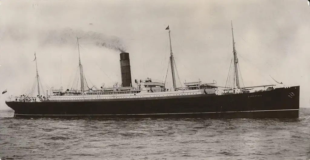 A vintage black and white photograph of a large steamship sailing on calm waters. The ship has a single towering smokestack and multiple masts. Smoke is billowing from the stack, and flags are visible on the masts.