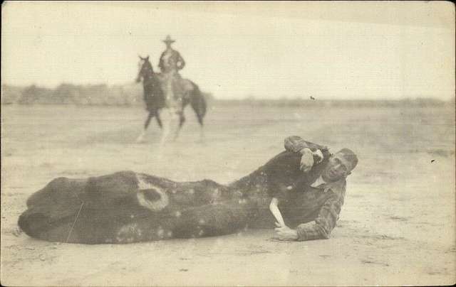 A cowboy lies on the ground playfully holding a calf. In the background, another cowboy sits on a horse, watching. The scene is set in an open, flat area.