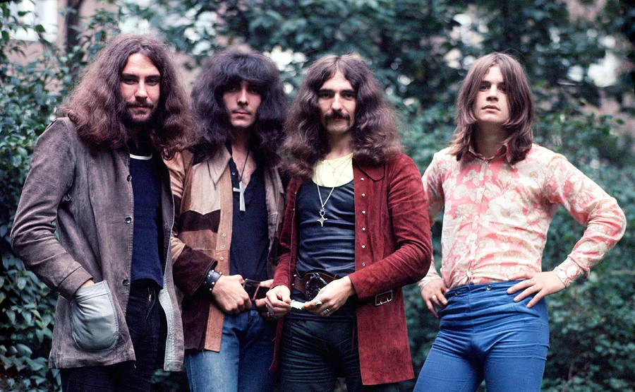 Four men with long hair stand outdoors in front of trees, wearing eclectic 1970s attire, including jackets, patterned shirts, and cross necklaces. They strike casual poses, exuding a laid-back, rock-and-roll vibe.