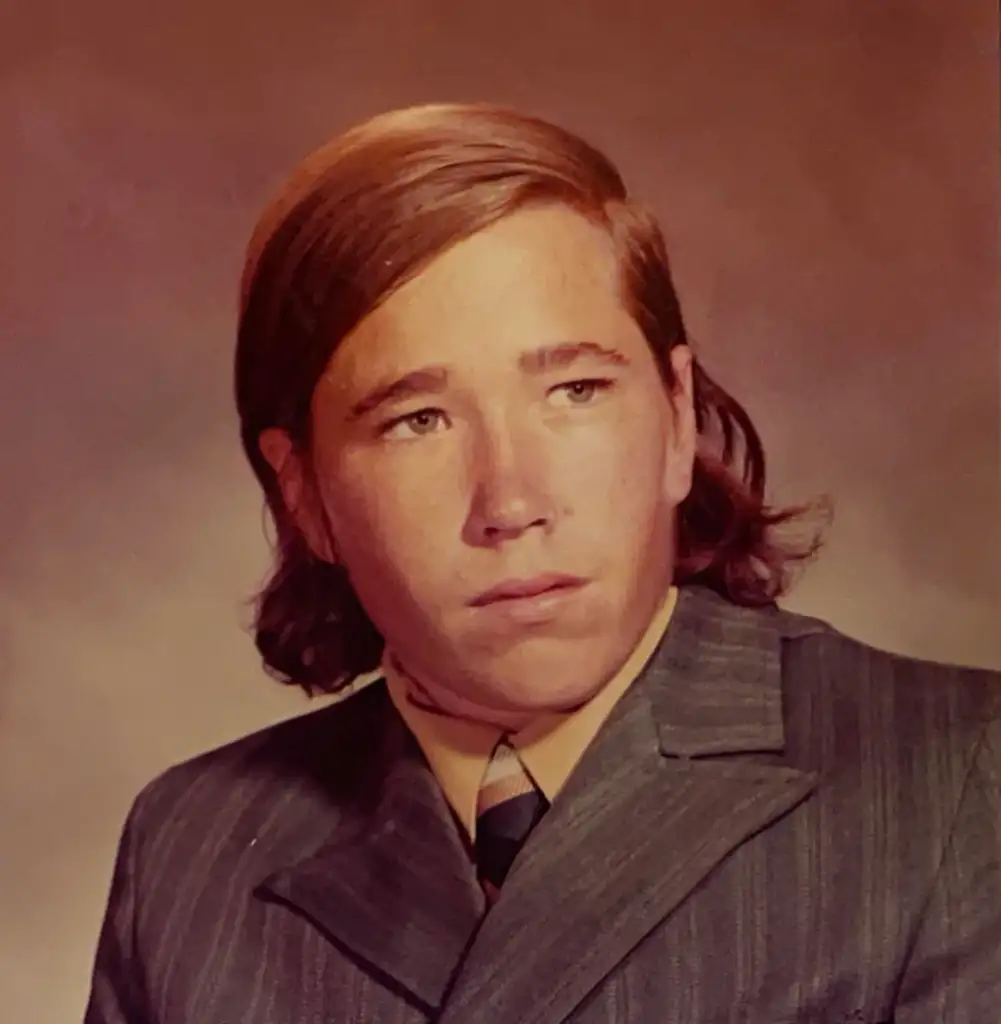 A young person with shoulder-length brown hair, wearing a striped suit and a light-colored shirt with a tie, poses for a formal portrait against a brown background.