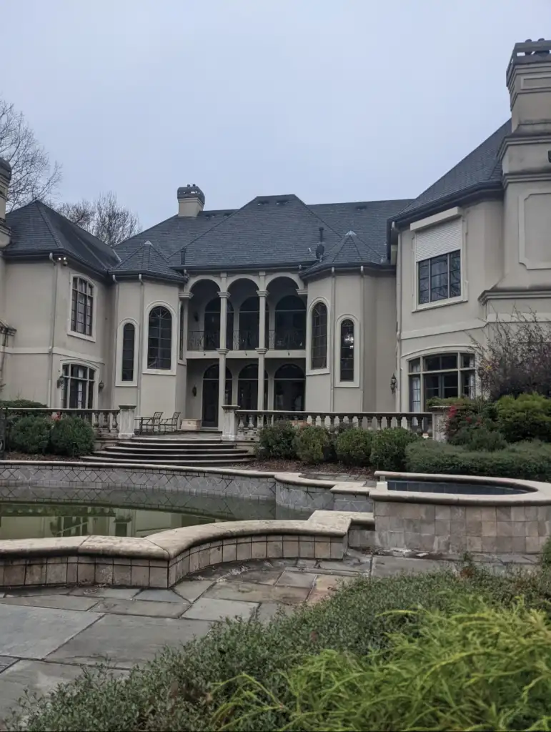 A large, elegant mansion with a gray roof and multiple arched windows. The backyard features a patio area and an empty pool. The surroundings are landscaped with shrubs and trees under an overcast sky.