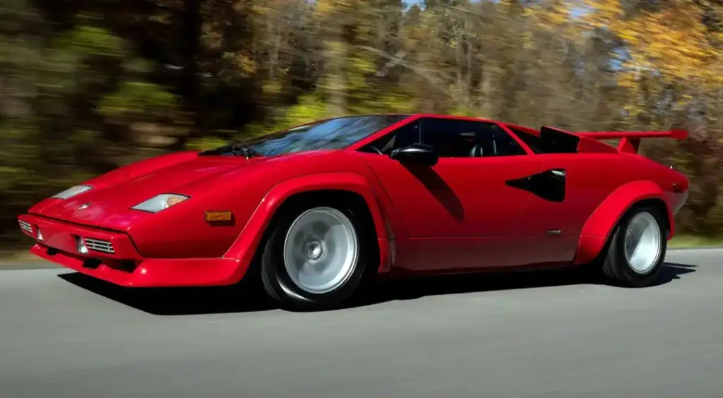 A red sports car, featuring an angular design and rear spoiler, drives on a road with blurred trees in the background, suggesting speed and motion on a sunny day.