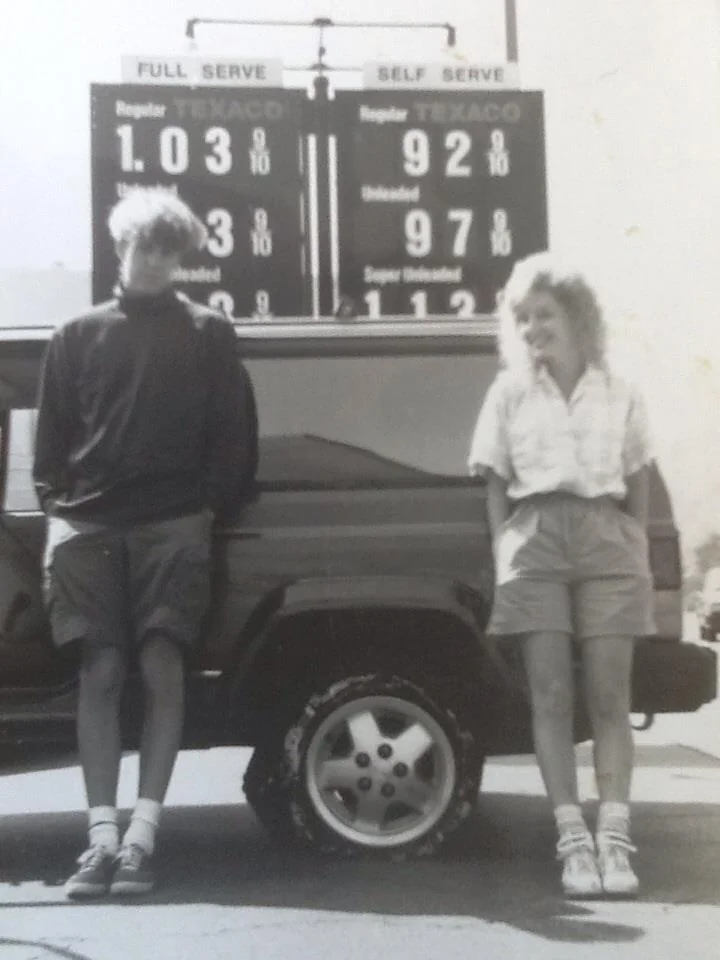 Two people stand leaning against a car in front of an old gas station sign displaying prices: Regular Full Serve at $1.03 and Self Serve at $0.92 per gallon. The photo is black and white, giving it a nostalgic feel.