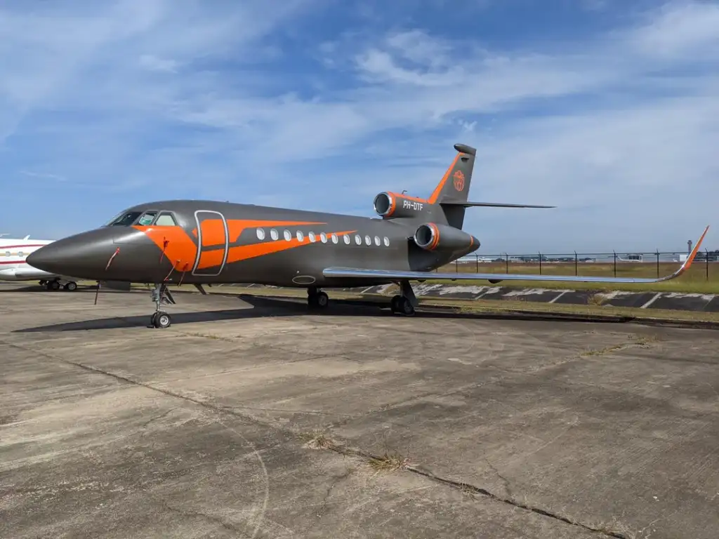 A sleek, gray private jet with orange accents is parked on an airport tarmac under a clear blue sky. The jet has a modern design, visible engines on the wings, and a tail section with an emblem. The ground is concrete with some grass in the background.