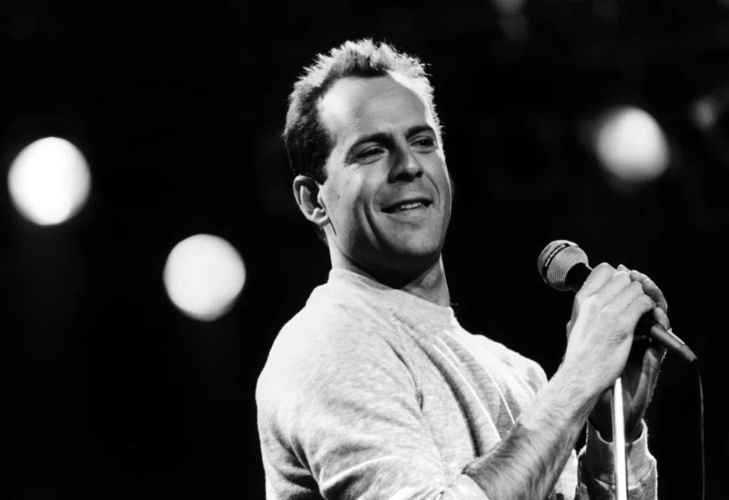 Black and white photo of a man with short hair, smiling while holding a microphone. He is wearing a light-colored sweater and stands against a blurred dark background with circular lights.