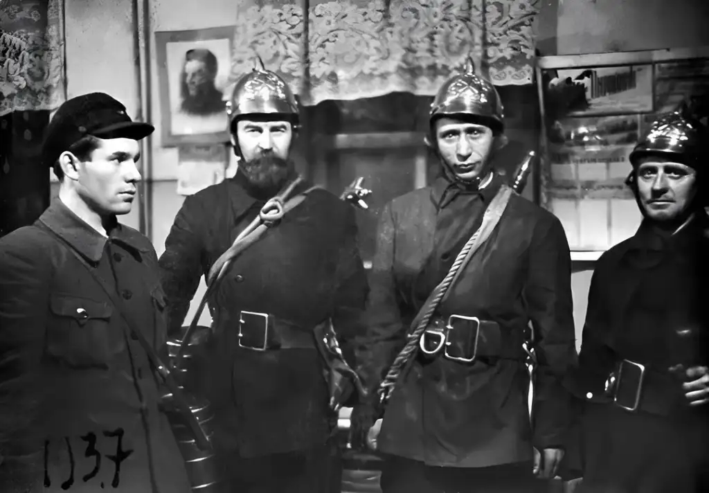 A black and white photo from 1937 shows four men dressed in vintage firefighter uniforms, including helmets and coats. They stand indoors against a backdrop of lace curtains and framed pictures on the wall.