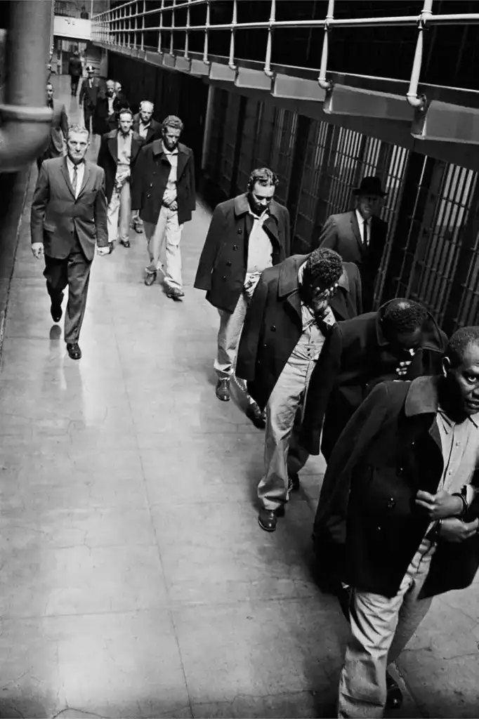 A group of inmates wearing prison uniforms and dark coats walk in a line through a prison corridor. They are escorted by guards. The corridor is lined with barred cells, and there is a balcony above. The scene is in black and white.