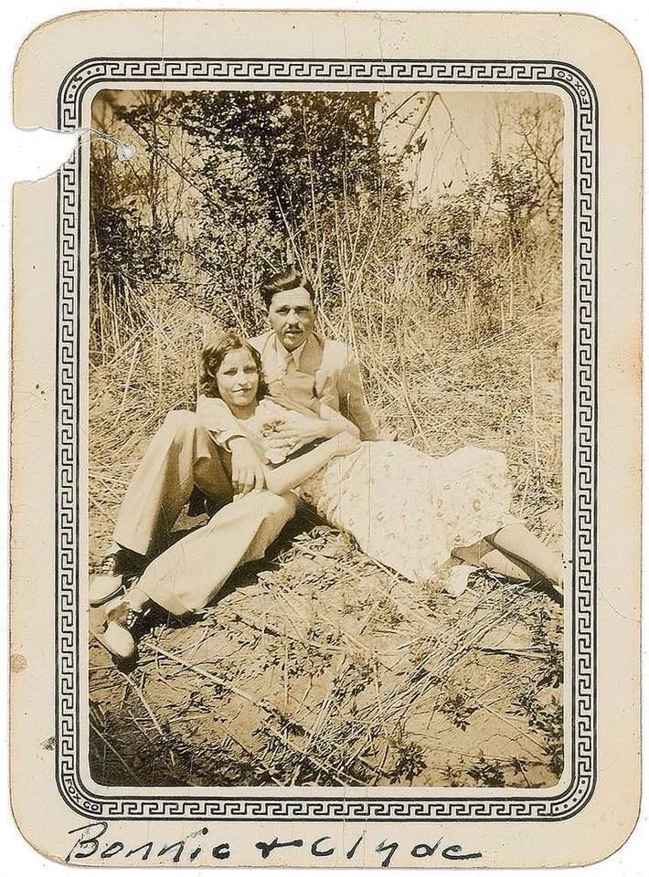 A sepia-toned vintage photo shows a couple sitting closely on the ground in an outdoor setting. The man is wearing a suit, and the woman is in a floral dress. The border has a decorative pattern, with "Bonnie & Clyde" handwritten at the bottom.