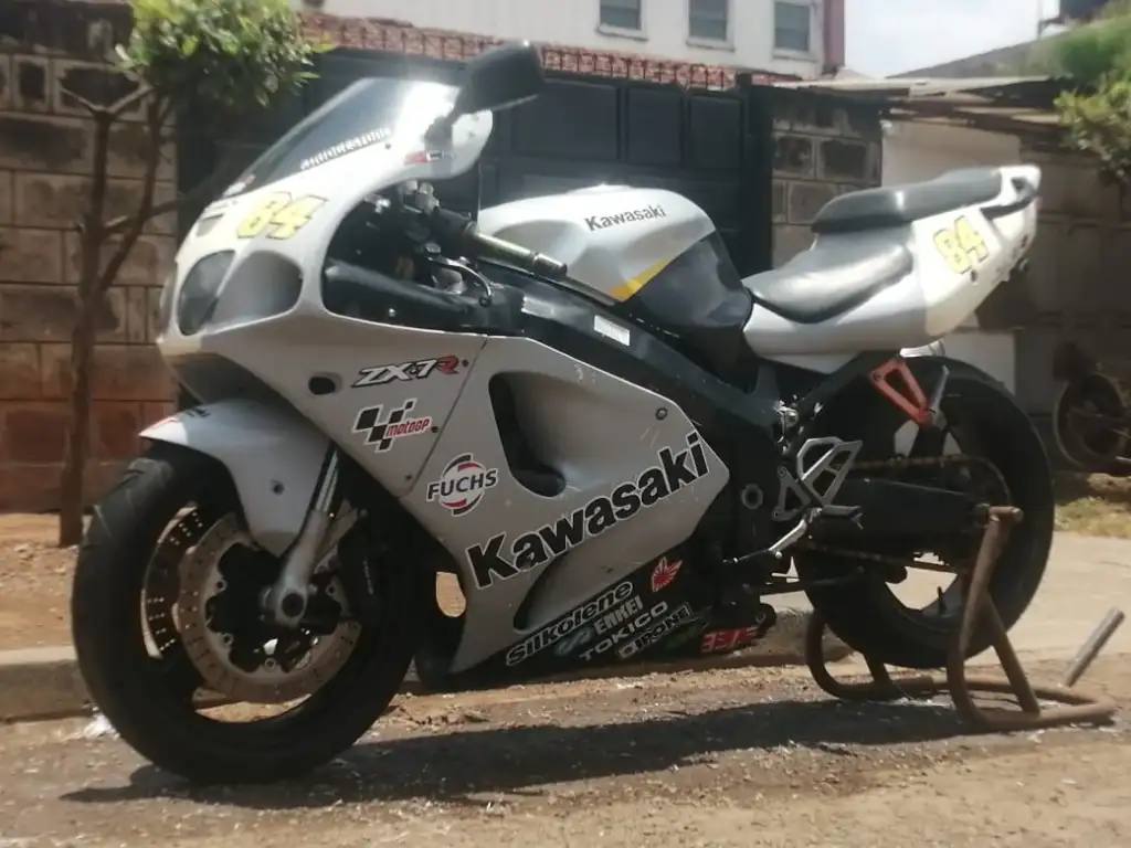 A white Kawasaki ZX-7R motorcycle with racing decals is parked on a stand on a sunny street. The bike features stickers, including "Silkolene," and the number 34 on the windshield and seat. The background shows a brick wall and greenery.