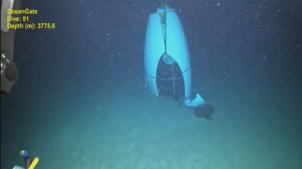 An underwater scene showing a large, capsule-like object resting on the ocean floor at a depth of 3775.6 meters. The water is dark, with particles floating around. Text indicates it's from OceanGate, Dive 01.