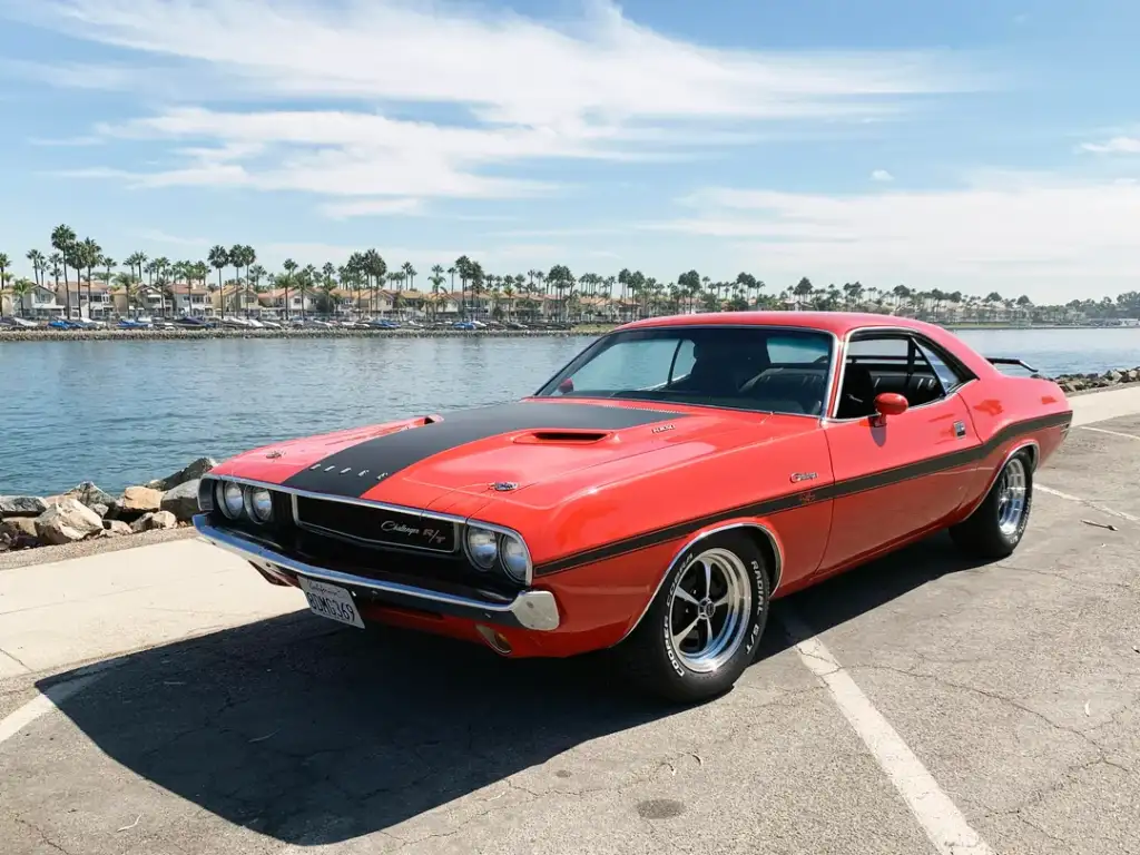 A classic red Dodge Challenger with a black hood is parked on a waterfront road. Palm trees and houses are visible in the background under a clear blue sky.