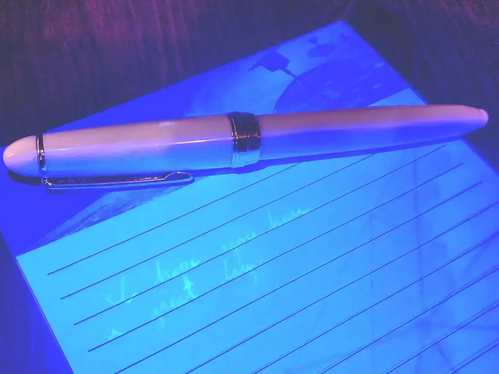 A white pen rests diagonally on a lined notepad, illuminated by blue and purple lighting. Some text is visible on the paper, though not clearly readable. The scene has a moody, colorful ambiance.