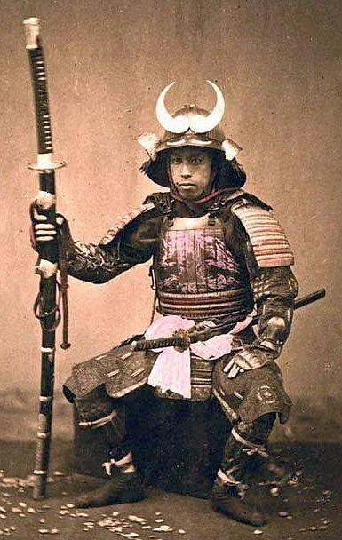 A person dressed in traditional samurai armor sits holding a long staff. The armor includes a helmet with prominent horns and ornate chest and shoulder pieces. The image is sepia-toned, suggesting a historical or vintage style.