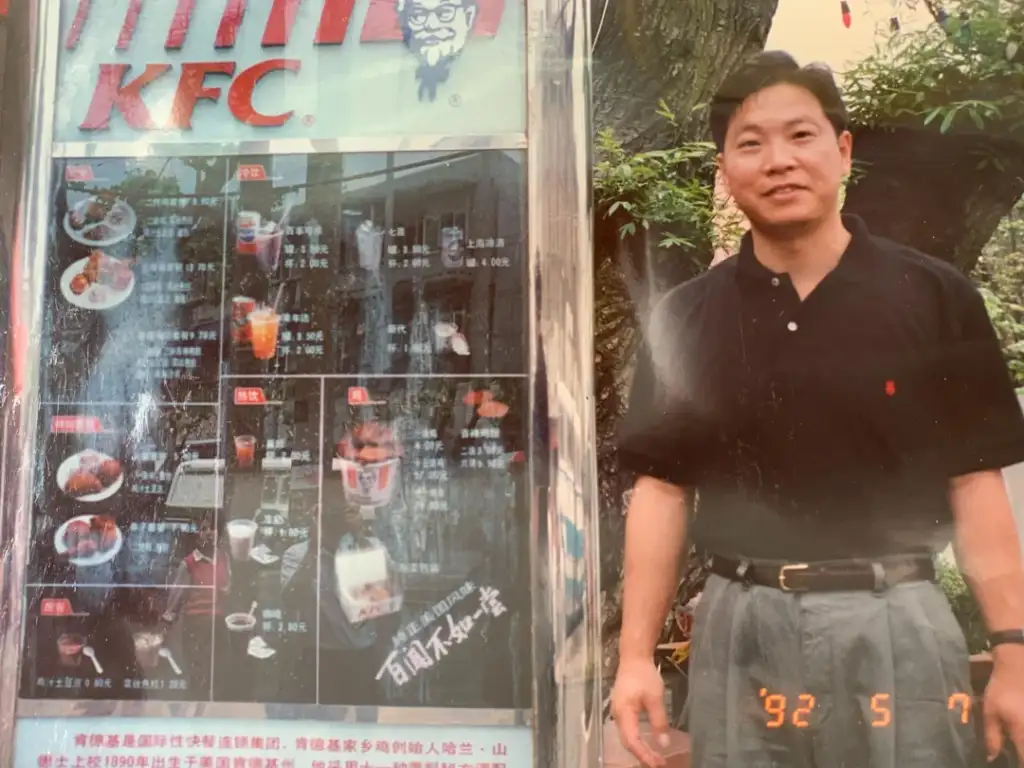 A man in a black polo and gray pants stands outdoors beside a KFC menu board displaying various food items and drinks. The image is slightly faded and dated May 7, 1992. Trees and greenery are visible in the background.